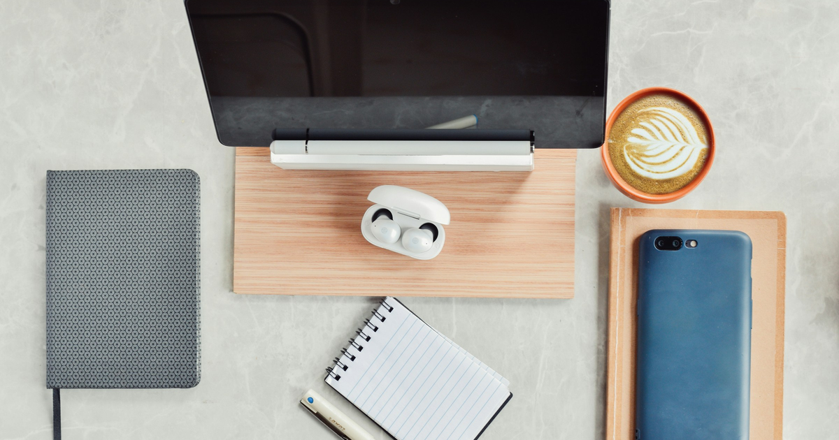 Overhead desk with a tablet, earbuds, notebooks, a phone, and a latte.