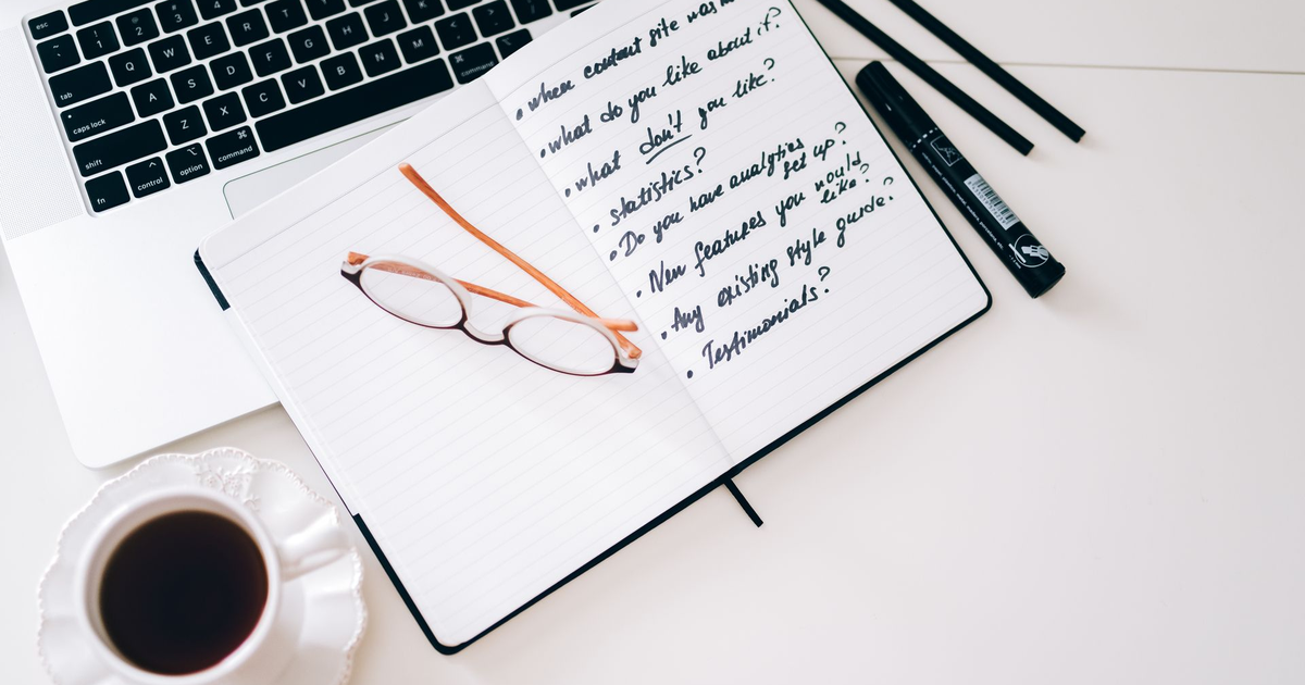 Open notebook with a handwritten list beside a laptop, glasses, and a cup of coffee on a white desk.